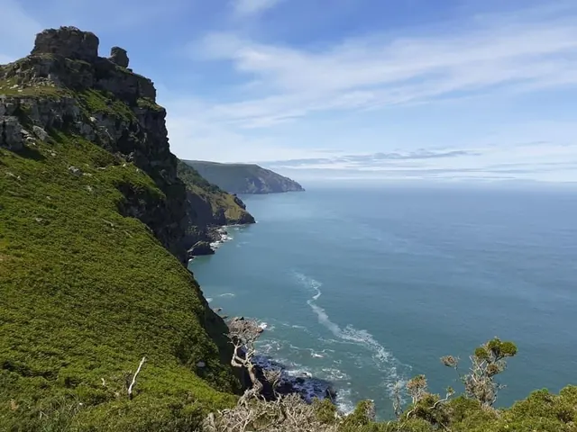 A craggy rock outcrop above a steep green slope, with the cliffs and headlands of the Exmoor coast running away into a calm blue sea