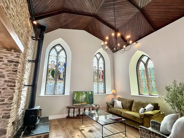 The nave of a converted chapel: two arched stained-glass windows, a wood-burner, a green velvet sofa under a timber roof