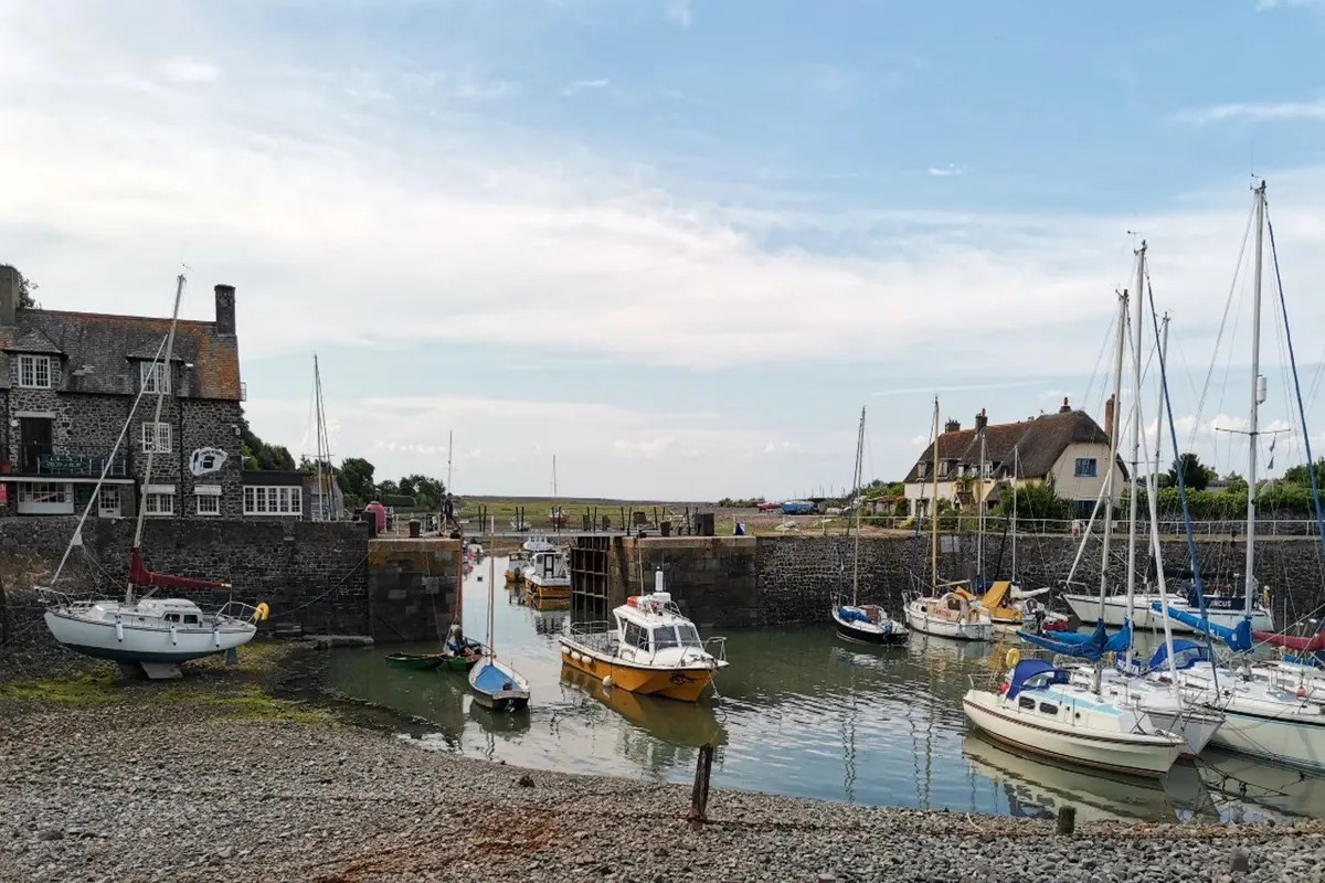 The inner harbour at Porlock Weir at low water, with yachts and a yellow-hulled launch moored below stone walls, a slate-roofed stone house on the left and a thatched cottage beyond the lock gates.